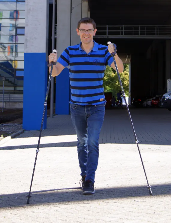 Ein Mann, der mit zwei Walking-Stöcken in Richtung Kamera läuft. Er hat eine Brille, ein Blau-Schwarz gestreiftes T-Sthirt und Bluejeans an.